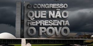 Imagem Acessível: A imagem mostra o prédio do Congresso Nacional em Brasília sob um céu nublado e escuro, com urubus voando e pousados no gramado em primeiro plano, transmitindo um clima de crítica e pessimismo. Em destaque, grandes letras de concreto rachadas trazem a frase: “O CONGRESSO QUE NÃO REPRESENTA O POVO”. Logo abaixo, sobre uma faixa branca e vermelha, repete-se a mensagem: “O Congresso que não representa o povo”. No canto superior esquerdo está escrito “PEC da Impunidade”, e no canto superior direito aparece o crédito da imagem “Fenajud”.