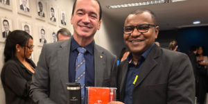 Imagem Acessível: A imagem mostra duas lideranças sorridentes posando lado a lado em um corredor do Congresso Nacional, com retratos em preto e branco de parlamentares ao fundo e outras pessoas circulando no ambiente. Ambos vestem paletó escuro; o homem à esquerda segura um copo térmico metálico, e o da direita segura uma agenda laranja com o logotipo da Fenajud. Na parte inferior da imagem, aparece o texto: “Fenajud reforça articulação no Congresso envolvendo Previdência e Reforma Administrativa”.