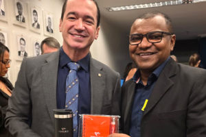 Imagem Acessível: A imagem mostra duas lideranças sorridentes posando lado a lado em um corredor do Congresso Nacional, com retratos em preto e branco de parlamentares ao fundo e outras pessoas circulando no ambiente. Ambos vestem paletó escuro; o homem à esquerda segura um copo térmico metálico, e o da direita segura uma agenda laranja com o logotipo da Fenajud. Na parte inferior da imagem, aparece o texto: “Fenajud reforça articulação no Congresso envolvendo Previdência e Reforma Administrativa”.