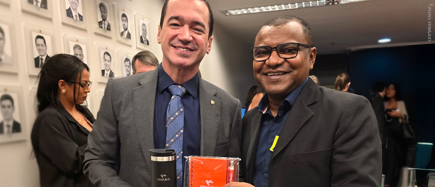 Imagem Acessível: A imagem mostra duas lideranças sorridentes posando lado a lado em um corredor do Congresso Nacional, com retratos em preto e branco de parlamentares ao fundo e outras pessoas circulando no ambiente. Ambos vestem paletó escuro; o homem à esquerda segura um copo térmico metálico, e o da direita segura uma agenda laranja com o logotipo da Fenajud. Na parte inferior da imagem, aparece o texto: “Fenajud reforça articulação no Congresso envolvendo Previdência e Reforma Administrativa”.