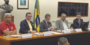 Imagem Acessível: A imagem mostra uma mesa de audiência pública na Câmara dos Deputados, com seis homens sentados lado a lado, entre parlamentares e representantes de entidades, tendo ao fundo a bandeira do Brasil. A maioria usa trajes formais. No rodapé da imagem está escrito: “Fenajud participa de audiência pública sobre superendividamento dos servidores públicos”.