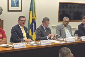 Imagem Acessível: A imagem mostra uma mesa de audiência pública na Câmara dos Deputados, com seis homens sentados lado a lado, entre parlamentares e representantes de entidades, tendo ao fundo a bandeira do Brasil. A maioria usa trajes formais. No rodapé da imagem está escrito: “Fenajud participa de audiência pública sobre superendividamento dos servidores públicos”.
