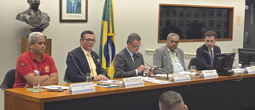 Imagem Acessível: A imagem mostra uma mesa de audiência pública na Câmara dos Deputados, com seis homens sentados lado a lado, entre parlamentares e representantes de entidades, tendo ao fundo a bandeira do Brasil. A maioria usa trajes formais. No rodapé da imagem está escrito: “Fenajud participa de audiência pública sobre superendividamento dos servidores públicos”.