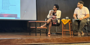 Cleonice Amorim de Paula, mulher negra integrante do SINJUS Antirracista, participa de uma mesa de debate sentada no palco, segurando um microfone enquanto observa a projeção de uma apresentação. Ao lado dela, um homem também participa da conversa, sentado com uma prancheta nas mãos. Ambos estão em cadeiras pretas, com uma mesa de centro entre eles e outro pequeno móvel com um tecido amarelo ao lado.