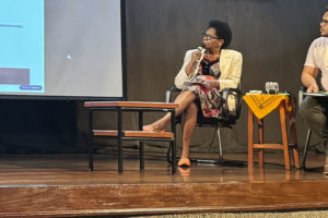 Cleonice Amorim de Paula, mulher negra integrante do SINJUS Antirracista, participa de uma mesa de debate sentada no palco, segurando um microfone enquanto observa a projeção de uma apresentação. Ao lado dela, um homem também participa da conversa, sentado com uma prancheta nas mãos. Ambos estão em cadeiras pretas, com uma mesa de centro entre eles e outro pequeno móvel com um tecido amarelo ao lado.