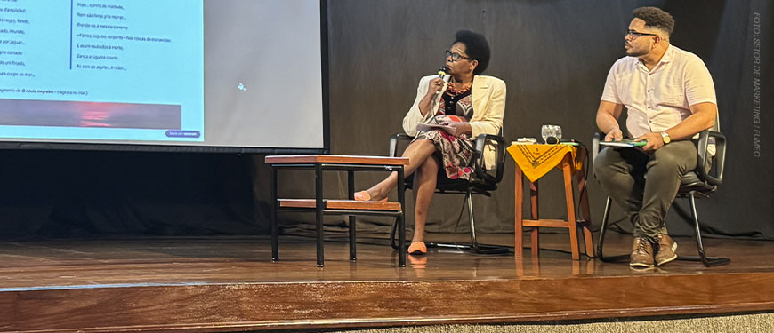 Cleonice Amorim de Paula, mulher negra integrante do SINJUS Antirracista, participa de uma mesa de debate sentada no palco, segurando um microfone enquanto observa a projeção de uma apresentação. Ao lado dela, um homem também participa da conversa, sentado com uma prancheta nas mãos. Ambos estão em cadeiras pretas, com uma mesa de centro entre eles e outro pequeno móvel com um tecido amarelo ao lado.
