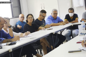 Imagem de uma reunião em uma mesa branca em formato circular com um grupo de aproximadamente quinze pessoas. Em primeiro plano há um notebook, um homem de óculos lendo um documento e ao fundo janelas abertas iluminando a sala com luz solar.