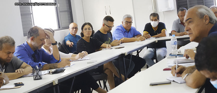 Imagem de uma reunião em uma mesa branca em formato circular com um grupo de aproximadamente quinze pessoas. Em primeiro plano há um notebook, um homem de óculos lendo um documento e ao fundo janelas abertas iluminando a sala com luz solar.