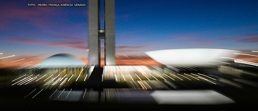 Imagem noturna do Congresso Nacional (duas torres centrais entre duas cúpulas, uma virada para baixo e outra virada para cima).