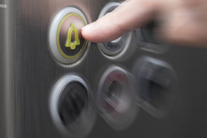 em destaque há a mão de uma pessoa acionando o botão de emergência no painel inox de um elevador.
