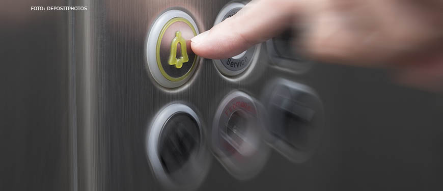em destaque há a mão de uma pessoa acionando o botão de emergência no painel inox de um elevador.