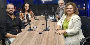 Imagem Acessível: Na mesa de um estúdio de gravação, há quatro pessoas sentadas: do lado esquerdo estão o coordenador-geral do SINJUS-MG, Felipe Rodrigues e a presidente da Associação Nacional dos Auditores Fiscais de Tributos Estaduais (Febrafite), Maria Aparecida Meloni (Papá), à direita estão Rose Laura Lopes Pinto, diretora da Associação dos Funcionários Fiscais de Minas Gerais (Affemg) e do Sindifisco-MG junto do anfitrião, o jornalista Orion Teixeira. Sobre a mesa há microfones, copos, papéis, um celular e um notebook. Ao fundo, uma televisão exibe o logotipo do podcast “Se Liga, Minas”, com o desenho do mapa de Minas Gerais e um ícone de podcast. Conteúdo textual: PODCAST - SINJUS debate educação fiscal em novo episódio do "Se Liga, Minas"
