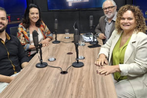 Imagem Acessível: Na mesa de um estúdio de gravação, há quatro pessoas sentadas: do lado esquerdo estão o coordenador-geral do SINJUS-MG, Felipe Rodrigues e a presidente da Associação Nacional dos Auditores Fiscais de Tributos Estaduais (Febrafite), Maria Aparecida Meloni (Papá), à direita estão Rose Laura Lopes Pinto, diretora da Associação dos Funcionários Fiscais de Minas Gerais (Affemg) e do Sindifisco-MG junto do anfitrião, o jornalista Orion Teixeira. Sobre a mesa há microfones, copos, papéis, um celular e um notebook. Ao fundo, uma televisão exibe o logotipo do podcast “Se Liga, Minas”, com o desenho do mapa de Minas Gerais e um ícone de podcast. Conteúdo textual: PODCAST - SINJUS debate educação fiscal em novo episódio do "Se Liga, Minas"