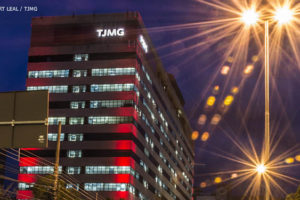 Vista noturna da sede do TJMG na Avenida Afonso Pena, há um efeito das luzes dos carros em movimento, formando linhas que seguem o contorno da avenida. Conteúdo textual: MESA DE NEGOCIAÇÃO - Sindicatos solicitam reunião com novo presidente do TJ.