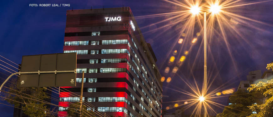 Vista noturna da sede do TJMG na Avenida Afonso Pena, há um efeito das luzes dos carros em movimento, formando linhas que seguem o contorno da avenida. Conteúdo textual: MESA DE NEGOCIAÇÃO - Sindicatos solicitam reunião com novo presidente do TJ.