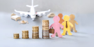 imagem com fundo em tons de cinza mostra desfocado um avião sobre uma mesa, acompanhado de pequenos objetos de viagem ao fundo como uma cama, um carro e um prato de comida. Em primeiro plano, há quatro pilhas de moedas em ordem crescente de altura. Ao lado das moedas, aparecem duas figuras de madeira coloridas representando pessoas: uma rosa segurando uma maleta e outra amarela usando gravata.