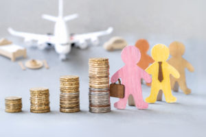 imagem com fundo em tons de cinza mostra desfocado um avião sobre uma mesa, acompanhado de pequenos objetos de viagem ao fundo como uma cama, um carro e um prato de comida. Em primeiro plano, há quatro pilhas de moedas em ordem crescente de altura. Ao lado das moedas, aparecem duas figuras de madeira coloridas representando pessoas: uma rosa segurando uma maleta e outra amarela usando gravata.