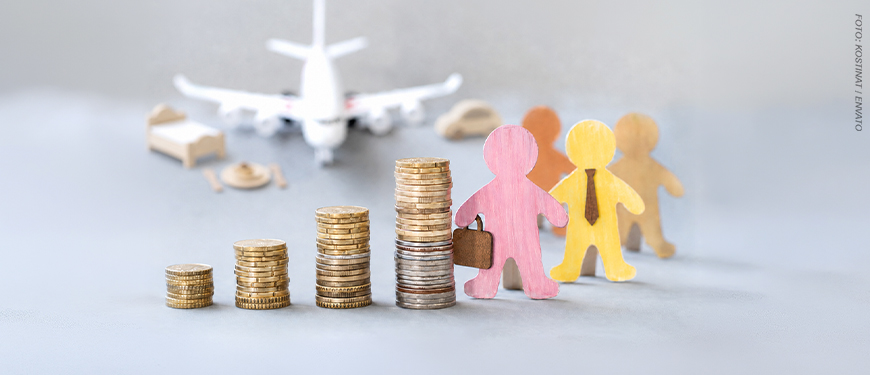 imagem com fundo em tons de cinza mostra desfocado um avião sobre uma mesa, acompanhado de pequenos objetos de viagem ao fundo como uma cama, um carro e um prato de comida. Em primeiro plano, há quatro pilhas de moedas em ordem crescente de altura. Ao lado das moedas, aparecem duas figuras de madeira coloridas representando pessoas: uma rosa segurando uma maleta e outra amarela usando gravata.