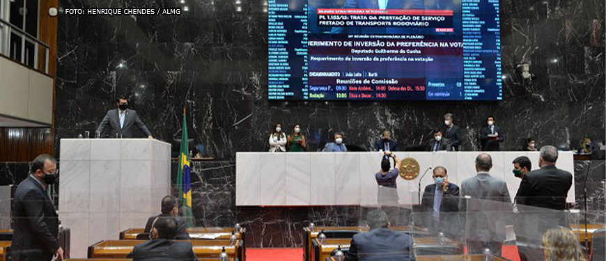 Deputados estaduais conversando na Mesa Diretora e no Plenário da Assembleia Legislativa de Minas Gerais. Ao fundo há uma parede em granito preto e um grande telão que projeta a imagem de um parlamentar que discursa na tribuna.