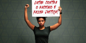 fundo cinza com destaque ao centro para um jovem negra, de cabelos crespos, usando camisa preta e segurando ao alto um cartaz com os dizeres: Lutar contra o racismo é fazer Justiça!