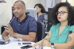 A imagem mostra um homem (Wagner Ferreira) e uma mulher (Iza Lourença) sentados à mesa lado a lado, o homem fala ao microfone em uma audiência na Câmara Municipal de BH. A mulher está vestindo uma camisa verde, é negra, de cabelos médios e cacheados, enquanto o homem está vestido com uma camisa azul, é negro e tem os cabelos grisalhos bem curtos.