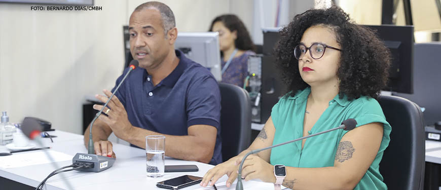 A imagem mostra um homem (Wagner Ferreira) e uma mulher (Iza Lourença) sentados à mesa lado a lado, o homem fala ao microfone em uma audiência na Câmara Municipal de BH. A mulher está vestindo uma camisa verde, é negra, de cabelos médios e cacheados, enquanto o homem está vestido com uma camisa azul, é negro e tem os cabelos grisalhos bem curtos.