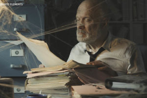 A imagem apresenta um homem mais velho, calvo e com barba branca, sentado em uma mesa em uma sala mal iluminada, com uma pilha de processos à sua frente. Ao seu redor há muitas teias de aranha, o ambiente aparenta estar sujo e desorganizado