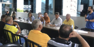 imagem de uma mesa diretiva composta de homens e mulheres durante uma reunião com destaque para o diretor de Finanças do SINJUS, Felipe Rodrigues (homem de pele clara, cabelo e barba castanhos, calça preta, camisa azul e óculos), que está de pé falando para a plateia.