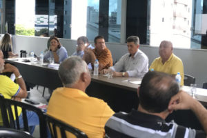 imagem de uma mesa diretiva composta de homens e mulheres durante uma reunião com destaque para o diretor de Finanças do SINJUS, Felipe Rodrigues (homem de pele clara, cabelo e barba castanhos, calça preta, camisa azul e óculos), que está de pé falando para a plateia.