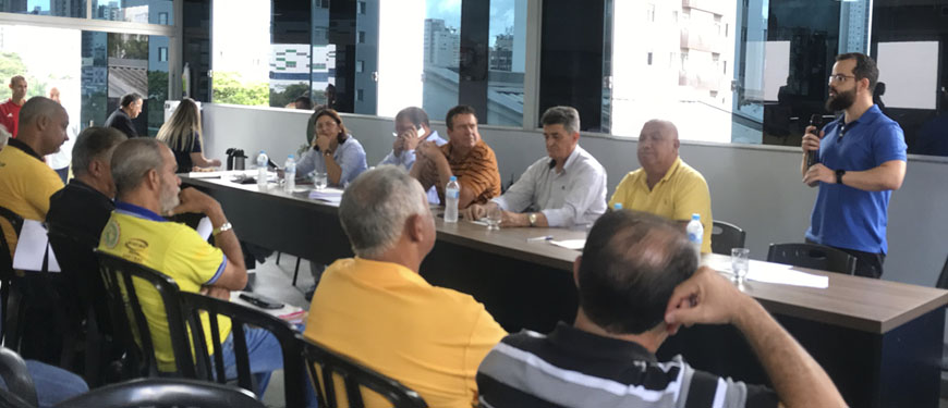 imagem de uma mesa diretiva composta de homens e mulheres durante uma reunião com destaque para o diretor de Finanças do SINJUS, Felipe Rodrigues (homem de pele clara, cabelo e barba castanhos, calça preta, camisa azul e óculos), que está de pé falando para a plateia.