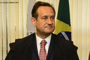 Fotografia do presidente eleito do TJMG, desembargador Luiz Carlos de Azevedo Corrêa Junior, ele está usando uma toga preta, com uma camisa branca e uma gravata vermelha com detalhes brancos. Ao fundo, há uma bandeira do Brasil e outra do estado de Minas Gerais