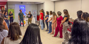 A imagem mostra um grupo de mulheres reunido durante uma atividade prática de defesa pessoal. Em destaque, o título diz “Workshop do NM ensina como identificar situações de risco e agir com segurança”. As participantes acompanham as orientações da instrutores durante a ação promovida pelo Núcleo das Mulheres (NM) do SINJUS-MG.