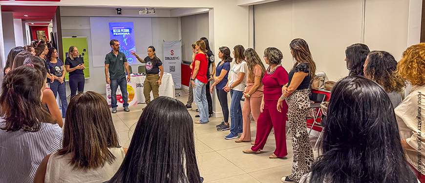 A imagem mostra um grupo de mulheres reunido durante uma atividade prática de defesa pessoal. Em destaque, o título diz “Workshop do NM ensina como identificar situações de risco e agir com segurança”. As participantes acompanham as orientações da instrutores durante a ação promovida pelo Núcleo das Mulheres (NM) do SINJUS-MG.