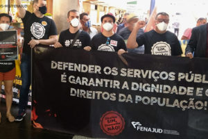 Servidores e servidoras, entre eles os diretores do SINJUS Alexandre Pires e Wagner Ferreira, protestando contra a Reforma Administrativa com cartazes e uma grande faixa preta com os dizeres na cor prata: “Defender os serviços públicos é garantir a dignidade e os direitos da população!”. A faixa conta com um selo dizendo “Direitos sim, desmonte não” e a logomarca da Fenajud.