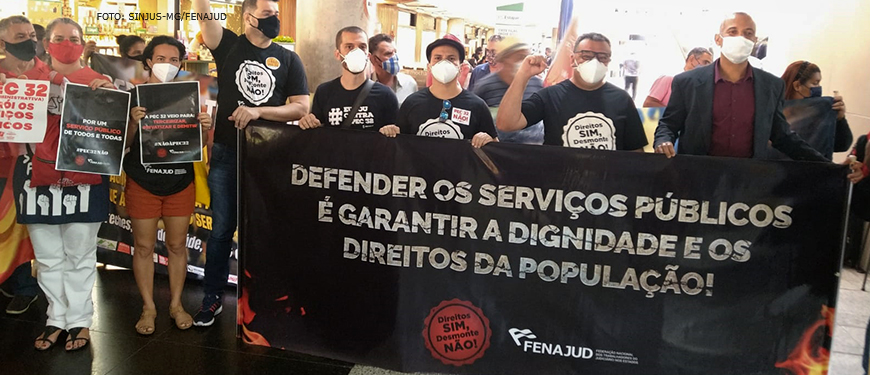 Servidores e servidoras, entre eles os diretores do SINJUS Alexandre Pires e Wagner Ferreira, protestando contra a Reforma Administrativa com cartazes e uma grande faixa preta com os dizeres na cor prata: “Defender os serviços públicos é garantir a dignidade e os direitos da população!”. A faixa conta com um selo dizendo “Direitos sim, desmonte não” e a logomarca da Fenajud.