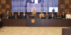 Fotografia em plano geral do auditório do TJMG durante a solenidade de abertura da Mostra de Tecnologias Assistivas. No palco, autoridades e representantes de pé e em posição de respeito perfilam-se atrás de uma longa bancada de madeira escura. Entre os integrantes da mesa e convidados estão Felipe Rodrigues (coordenador-geral do SINJUS), Alex Aguiar (coordenador do Núcleo da Pessoa com Deficiência - NPD), o desembargador Manoel dos Reis Morais (2º vice-presidente do TJMG), Raimundo Messias Júnior (Corregedor-geral de Justiça), Eduardo Couto (presidente do SERJUSMIG), Roberto Carlos de Arruda (diretor do SINDOJUS/MG), além de representantes da magistratura e dos ministérios da Ciência e Tecnologia e dos Direitos Humanos e Cidadania.