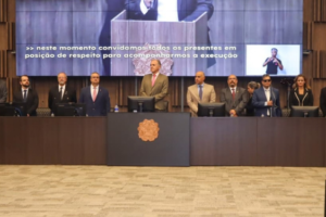 Fotografia em plano geral do auditório do TJMG durante a solenidade de abertura da Mostra de Tecnologias Assistivas. No palco, autoridades e representantes de pé e em posição de respeito perfilam-se atrás de uma longa bancada de madeira escura. Entre os integrantes da mesa e convidados estão Felipe Rodrigues (coordenador-geral do SINJUS), Alex Aguiar (coordenador do Núcleo da Pessoa com Deficiência - NPD), o desembargador Manoel dos Reis Morais (2º vice-presidente do TJMG), Raimundo Messias Júnior (Corregedor-geral de Justiça), Eduardo Couto (presidente do SERJUSMIG), Roberto Carlos de Arruda (diretor do SINDOJUS/MG), além de representantes da magistratura e dos ministérios da Ciência e Tecnologia e dos Direitos Humanos e Cidadania.