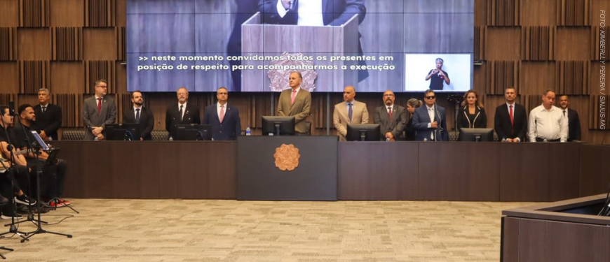 Fotografia em plano geral do auditório do TJMG durante a solenidade de abertura da Mostra de Tecnologias Assistivas. No palco, autoridades e representantes de pé e em posição de respeito perfilam-se atrás de uma longa bancada de madeira escura. Entre os integrantes da mesa e convidados estão Felipe Rodrigues (coordenador-geral do SINJUS), Alex Aguiar (coordenador do Núcleo da Pessoa com Deficiência - NPD), o desembargador Manoel dos Reis Morais (2º vice-presidente do TJMG), Raimundo Messias Júnior (Corregedor-geral de Justiça), Eduardo Couto (presidente do SERJUSMIG), Roberto Carlos de Arruda (diretor do SINDOJUS/MG), além de representantes da magistratura e dos ministérios da Ciência e Tecnologia e dos Direitos Humanos e Cidadania.