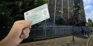 Imagem Acessível: A imagem mostra uma mão segurando uma carteira funcional do Tribunal de Justiça de Minas Gerais, diante do prédio da unidade Milton Campos, em Belo Horizonte. O texto na imagem informa: “Posto temporário para emissão da carteira funcional está aberto na unidade Milton Campos”.