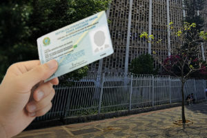 Imagem Acessível: A imagem mostra uma mão segurando uma carteira funcional do Tribunal de Justiça de Minas Gerais, diante do prédio da unidade Milton Campos, em Belo Horizonte. O texto na imagem informa: “Posto temporário para emissão da carteira funcional está aberto na unidade Milton Campos”.