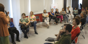 Imagem Acessível: A imagem mostra uma sala ampla e iluminada, onde a jornalista Paula Bento fala ao microfone diante de um grupo de aposentadas e aposentados do SINJUS-MG, durante o encontro do NAP, que refletiu sobre o uso de telas e bem-estar dos aposentados. Ao fundo, há um telão com a apresentação e um banner do Núcleo.