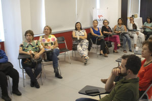 Imagem Acessível: A imagem mostra uma sala ampla e iluminada, onde a jornalista Paula Bento fala ao microfone diante de um grupo de aposentadas e aposentados do SINJUS-MG, durante o encontro do NAP, que refletiu sobre o uso de telas e bem-estar dos aposentados. Ao fundo, há um telão com a apresentação e um banner do Núcleo.