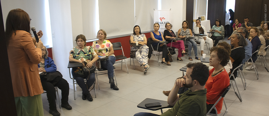 Imagem Acessível: A imagem mostra uma sala ampla e iluminada, onde a jornalista Paula Bento fala ao microfone diante de um grupo de aposentadas e aposentados do SINJUS-MG, durante o encontro do NAP, que refletiu sobre o uso de telas e bem-estar dos aposentados. Ao fundo, há um telão com a apresentação e um banner do Núcleo.