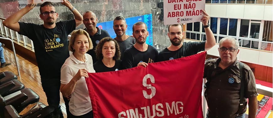 Foto dos diretores do SINJUS-MG segurando a bandeira do Sinjus na cor vermelha e cartazes com os dizeres "DATA-BASE EU NÃO ABRO MÃO!" no plenário da ALMG.