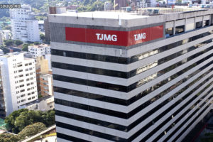 Na imagem estão os últimos andares do edifício sede do Tribunal de Justiça de Minas Gerais com um letreiro vermelho onde está escrito TJMG em branco. Ao fundo, estão uma parte da Serra do Curral com vegetação verde e edifícios vizinhos ao Tribunal. Conteúdo textual: ATUALIZAÇÃO - Carteira funcional do TJMG: agendamento para coleta de dados está disponível.