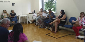 Fotografia de uma sala de reuniões com paredes brancas e chão de taco, onde se vê pessoas sentadas em carteiras debatendo.