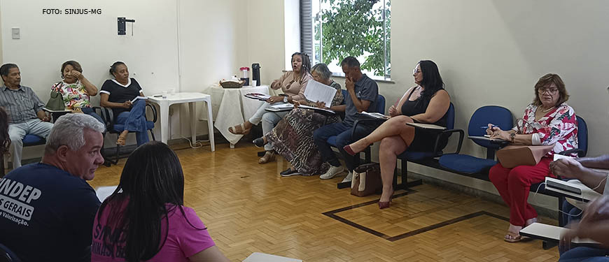 Fotografia de uma sala de reuniões com paredes brancas e chão de taco, onde se vê pessoas sentadas em carteiras debatendo.