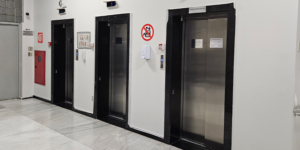 A imagem mostra um corredor do Anexo 1, com quatro elevadores lado a lado, todos de portas metálicas e moldura preta. Acima de cada elevador há um visor digital azul indicando o andar em que cada cabine está.