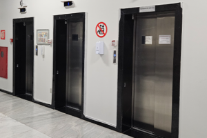 A imagem mostra um corredor do Anexo 1, com quatro elevadores lado a lado, todos de portas metálicas e moldura preta. Acima de cada elevador há um visor digital azul indicando o andar em que cada cabine está.