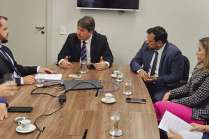 Fotografia de uma reunião realizada em uma sala com paredes claras e uma mesa retangular de madeira. Há oito pessoas estão sentadas ao redor da mesa. No centro da imagem, um homem de terno preto e gravata listrada fala gesticulando com as mãos, enquanto outros participantes ouvem atentamente e tomam notas.
